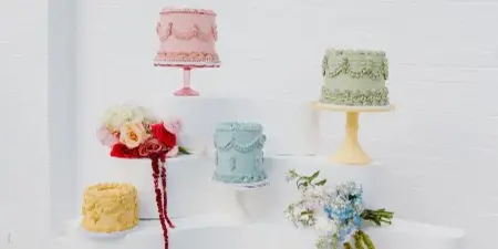 A display of various custom cakes and pastries arranged on a white tiered stand against a white brick wall.