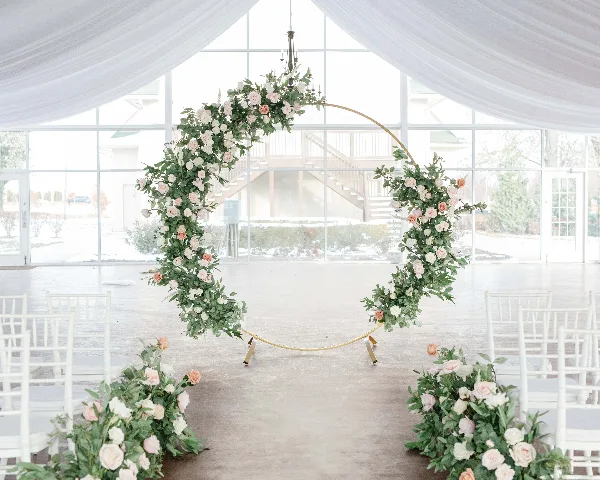 A gold wedding arch decorated with white and pink flowers and greenery, set up in a venue with white Chiavari chairs.