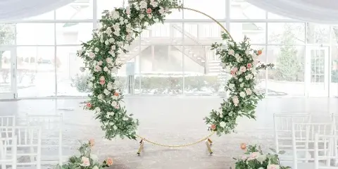 A gold wedding arch decorated with white and pink flowers and greenery, set up in a venue with white Chiavari chairs.