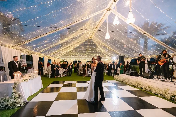 A bride and groom dancing on a black and white checkered floor inside a large wedding tent decorated with string lights and white floral arrangements.