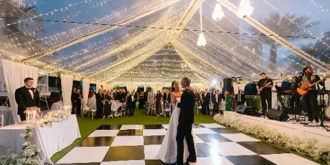 A bride and groom dancing on a black and white checkered floor inside a large wedding tent decorated with string lights and white floral arrangements.