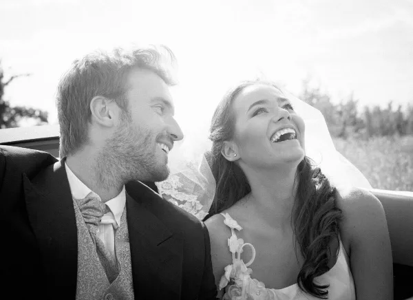 A black and white photograph of a bride and groom laughing together while sitting in a car.