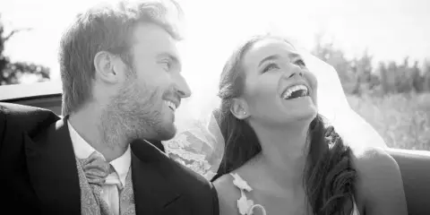 A black and white photograph of a bride and groom laughing together while sitting in a car.