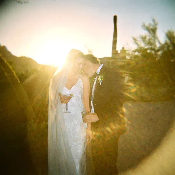 A bride and groom sharing a moment outdoors at sunset, with the bride holding a cocktail and the groom kissing her cheek.
