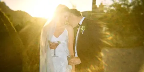 A bride and groom sharing a moment outdoors at sunset, with the bride holding a cocktail and the groom kissing her cheek.