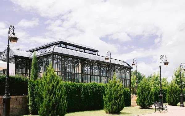Exterior view of a large glass conservatory building with iron framing, surrounded by green hedges and decorative lamp posts.