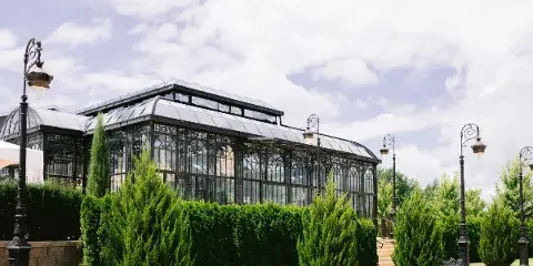 Exterior view of a large glass conservatory building with iron framing, surrounded by green hedges and decorative lamp posts.