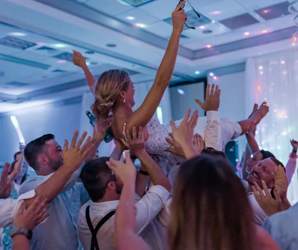 A bride in a white lace dress is lifted into the air by a crowd of cheering wedding guests in a banquet hall.