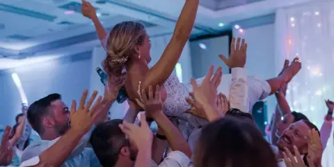A bride in a white lace dress is lifted into the air by a crowd of cheering wedding guests in a banquet hall.