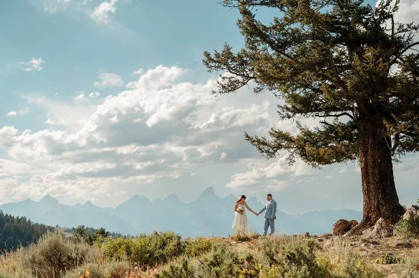 A bride and groom holding hands on a grassy hillside with the Teton mountain range in the background.