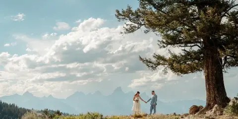 A bride and groom holding hands on a grassy hillside with the Teton mountain range in the background.