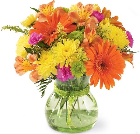 A colorful mixed bouquet of fresh flowers including orange gerbera daisies, yellow chrysanthemums, and pink carnations in a green glass vase.