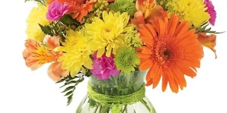 A colorful mixed bouquet of fresh flowers including orange gerbera daisies, yellow chrysanthemums, and pink carnations in a green glass vase.