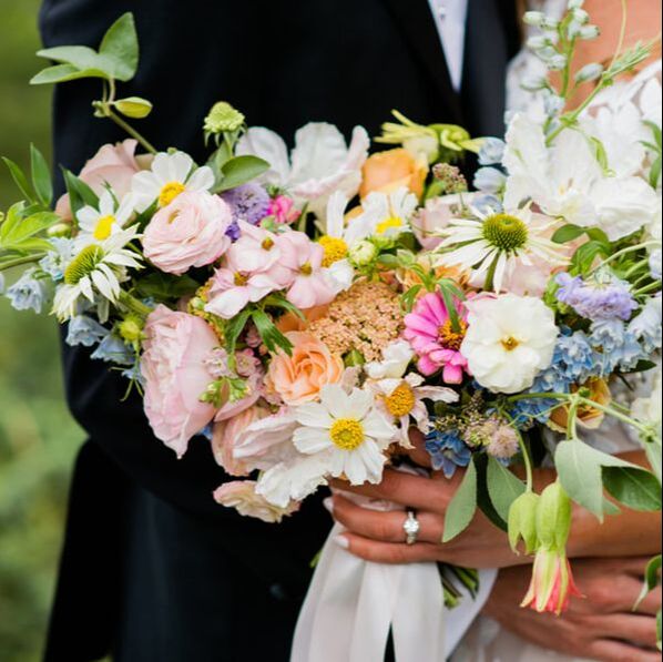 A close-up photograph of a colorful, lush bridal bouquet featuring roses, daisies, and greenery held by a couple.
