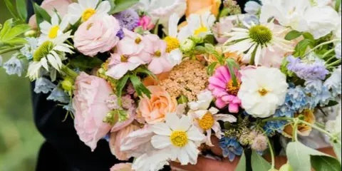 A close-up photograph of a colorful, lush bridal bouquet featuring roses, daisies, and greenery held by a couple.