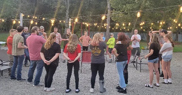 A group of people standing in a circle on a gravel outdoor patio with string lights overhead and a white fence in the background.