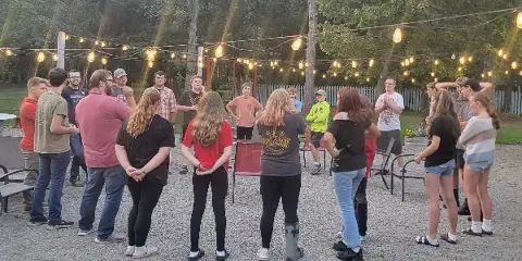 A group of people standing in a circle on a gravel outdoor patio with string lights overhead and a white fence in the background.