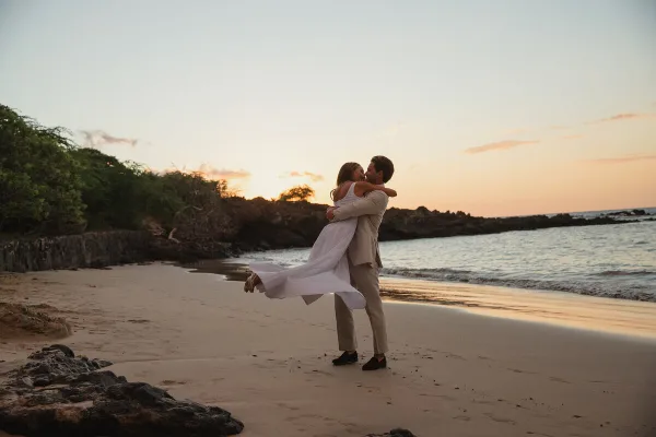 A wedding couple embracing on a sandy beach at sunset, with the bride's white dress flowing in the wind.