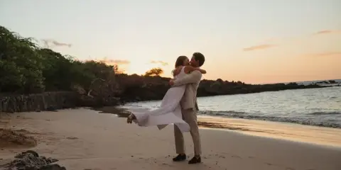 A wedding couple embracing on a sandy beach at sunset, with the bride's white dress flowing in the wind.