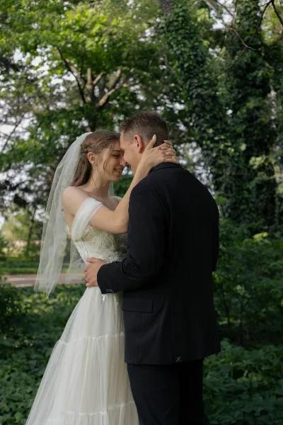 A bride in a white gown and veil and a groom in a black suit embracing and smiling in a garden setting.