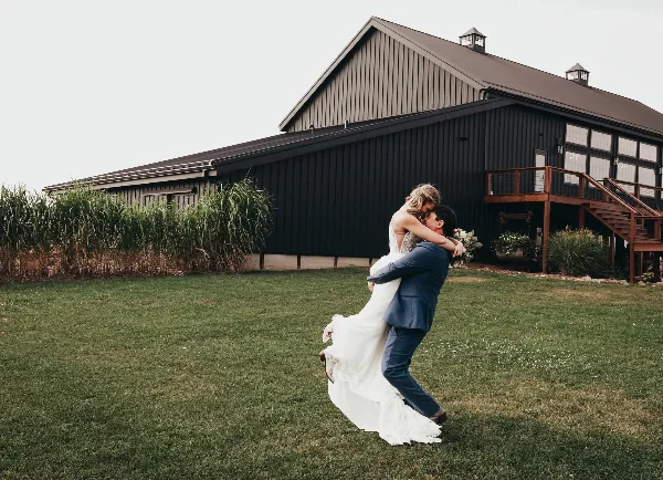 A bride and groom dancing on a lawn in front of a large, dark-colored event barn.