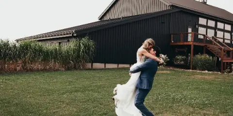 A bride and groom dancing on a lawn in front of a large, dark-colored event barn.