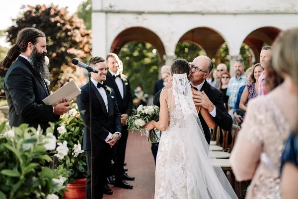 A bride and groom sharing a kiss during an outdoor wedding ceremony with an officiant and guests present.