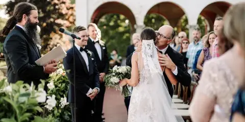 A bride and groom sharing a kiss during an outdoor wedding ceremony with an officiant and guests present.