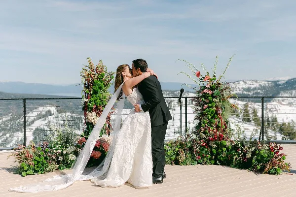 A bride and groom kissing in front of a large floral arch and floor arrangements on a mountain terrace.