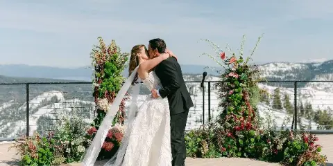 A bride and groom kissing in front of a large floral arch and floor arrangements on a mountain terrace.