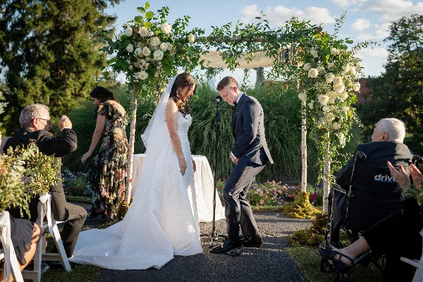A bride and groom standing under a floral arch decorated with white roses and greenery during an outdoor wedding ceremony.