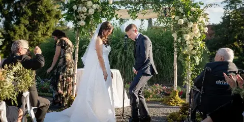 A bride and groom standing under a floral arch decorated with white roses and greenery during an outdoor wedding ceremony.