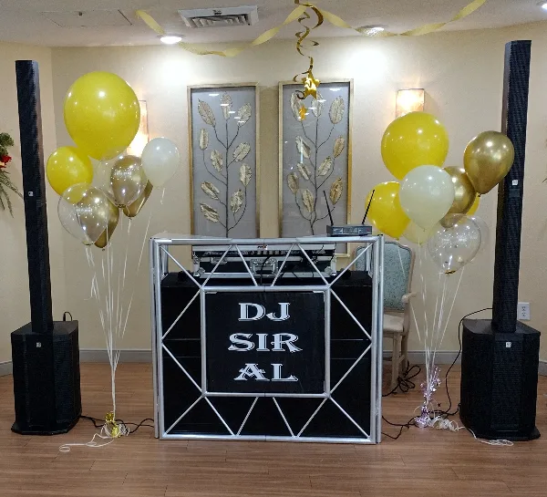 A professional DJ booth setup featuring a 'DJ SIR AL' branded backdrop, large speakers, and yellow and gold balloons in an event venue.