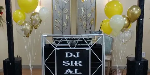 A professional DJ booth setup featuring a 'DJ SIR AL' branded backdrop, large speakers, and yellow and gold balloons in an event venue.