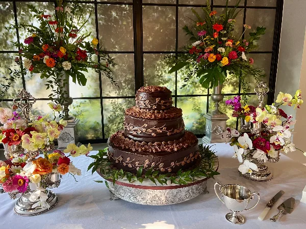 A three-tiered chocolate wedding cake with floral decorations on a silver stand, surrounded by flower arrangements.