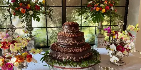 A three-tiered chocolate wedding cake with floral decorations on a silver stand, surrounded by flower arrangements.