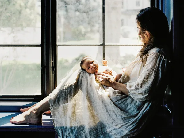 A mother sitting by a window holding a newborn baby.