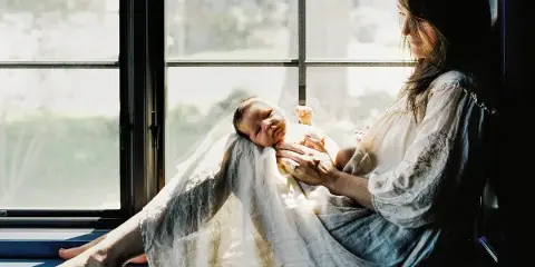 A mother sitting by a window holding a newborn baby.