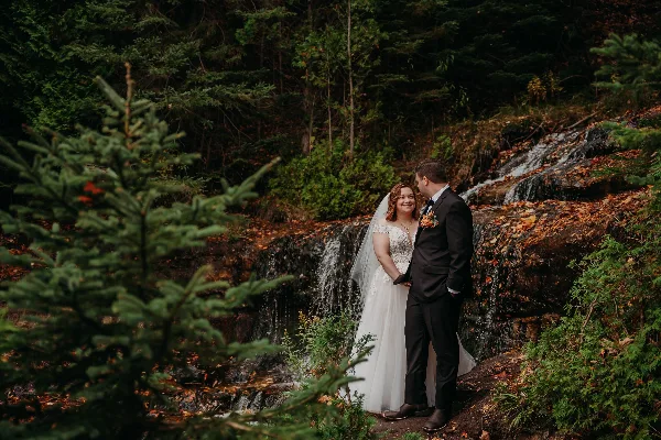A bride and groom standing together in a forest near a small waterfall.