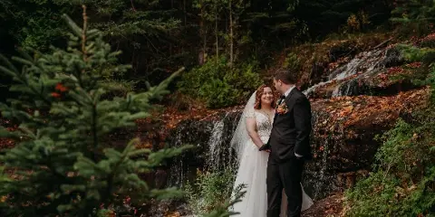 A bride and groom standing together in a forest near a small waterfall.