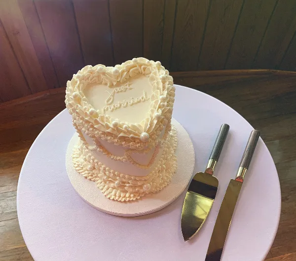 A heart-shaped, two-tiered white wedding cake decorated with intricate buttercream piping and pearls, featuring the words 'Just Married' on the top tier, sitting on a white cake board next to a gold cake server and knife.