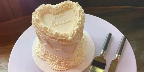A heart-shaped, two-tiered white wedding cake decorated with intricate buttercream piping and pearls, featuring the words 'Just Married' on the top tier, sitting on a white cake board next to a gold cake server and knife.