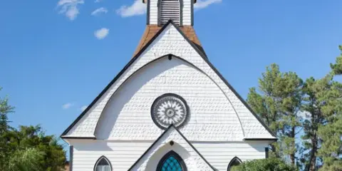 Exterior view of a white historic chapel with a steeple and cross under a blue sky.