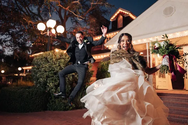 A bride and groom jumping joyfully in front of a venue at night, with the bride holding a bouquet.