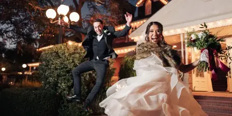 A bride and groom jumping joyfully in front of a venue at night, with the bride holding a bouquet.