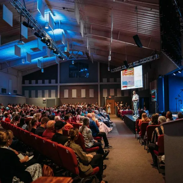 Interior view of a large church auditorium with red seating, a stage, and a congregation attending a service.