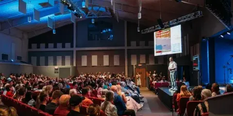 Interior view of a large church auditorium with red seating, a stage, and a congregation attending a service.