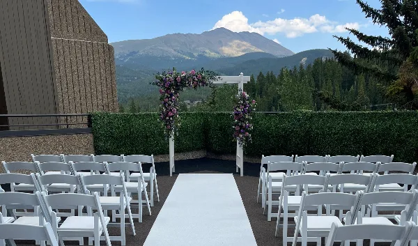 A wedding ceremony setup with a floral arch and white chairs on a deck overlooking a mountain range.