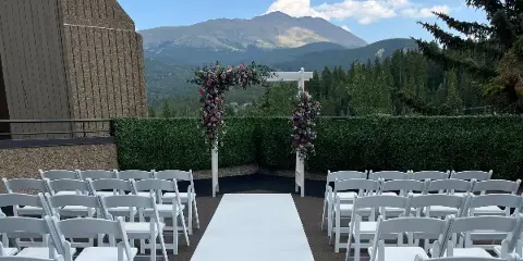 A wedding ceremony setup with a floral arch and white chairs on a deck overlooking a mountain range.