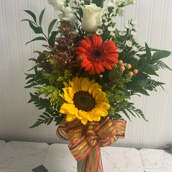 A tall mixed floral bouquet with sunflowers, a red gerbera daisy, and white blooms in a glass vase.